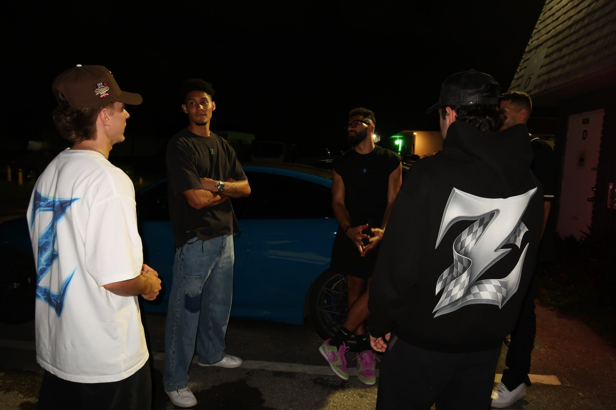 Group of young men (Gen Z) hanging out at night beside a blue BMW, wearing Genzerz streetwear. One wears the Racer Z hoodie with the signature chrome Z logo on the back. The scene captures Genzerz culture motion, freedom, and unity.