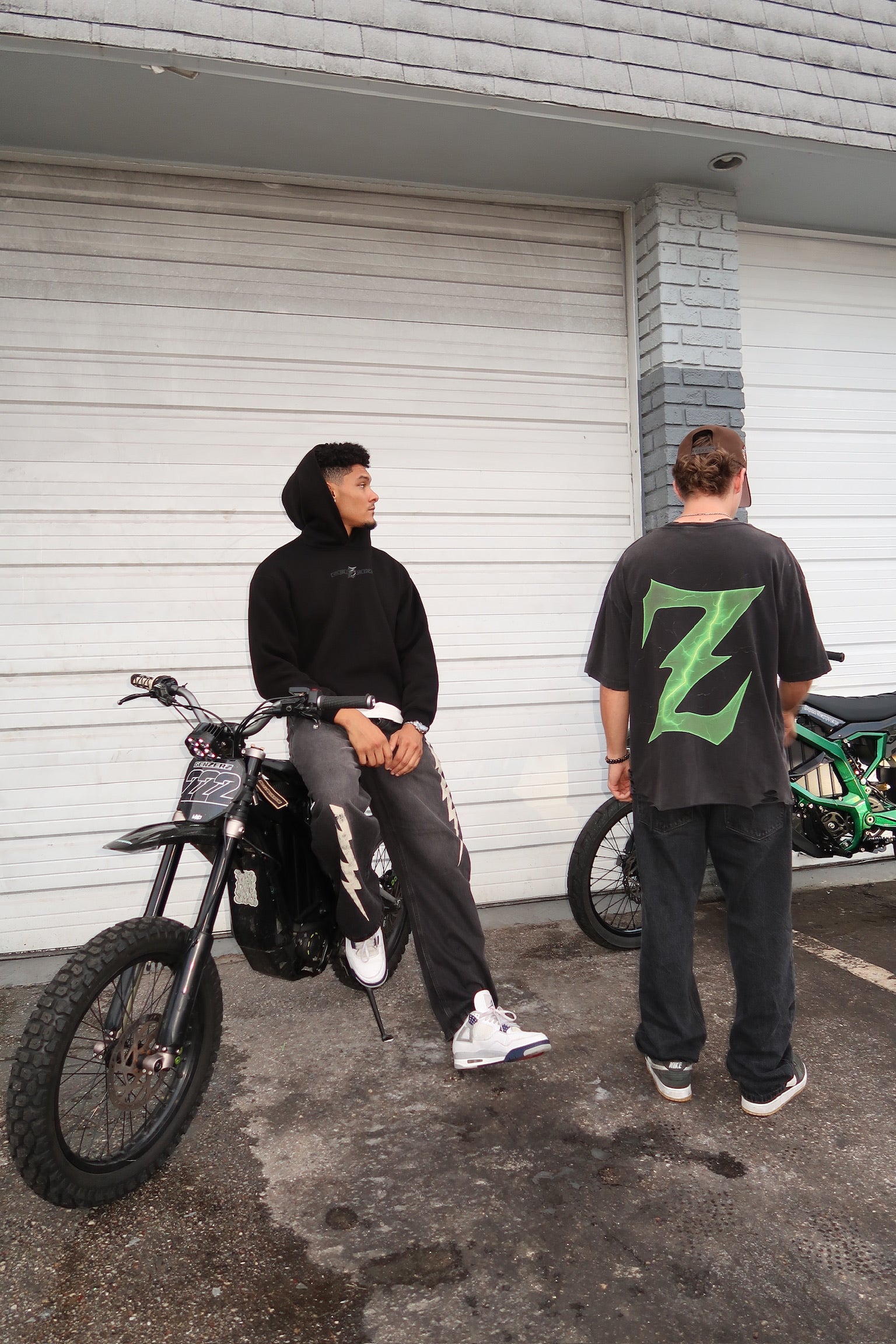 Two riders wearing Genzerz streetwear Standing beside Sur-Ron e-bikes outside a closed garage. The scene captures the brand’s underground street culture and motion-driven lifestyle.
