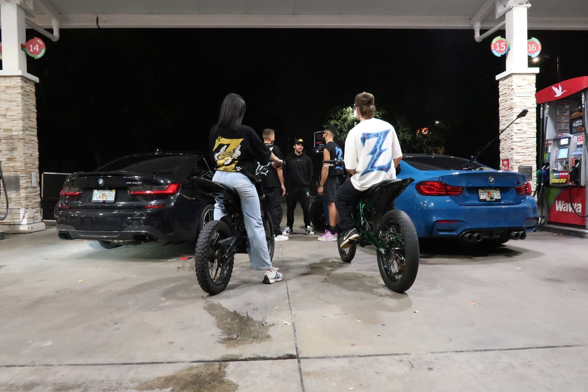 Two riders on Sur-Ron e-bikes wearing Genzerz streetwear, parked at a gas station beside BMWs at night. The scene captures freedom, adrenaline, and the motion-driven culture behind Genzerz Co.