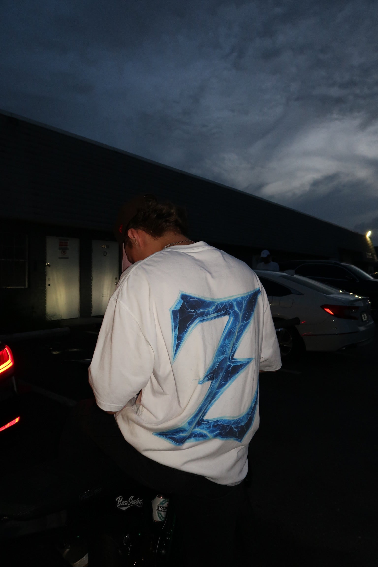 Person wearing a white Genzerz hoodie with a large blue Z logo on the back, standing near cars at night. The photo captures the brand’s signature streetwear style and motion-driven culture.